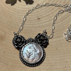 K&ouml;nnte beinhalten: Eine Silberkette mit einer zentralen, runden, schillernden Perle in einer Fassung aus dunkel oxidiertem Silber. Der Anh&auml;nger wird von zwei detaillierten silbernen Blumenmotiven und einer kleinen zentralen Blume flankiert, an einer feinen ovalen Gliederkette.