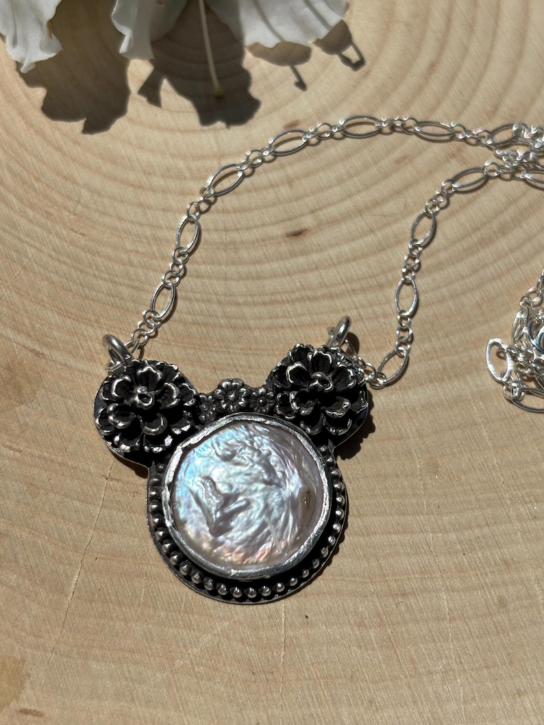 K&ouml;nnte beinhalten: Eine Silberkette mit einer zentralen, runden, schillernden Perle in einer Fassung aus dunkel oxidiertem Silber. Der Anh&auml;nger wird von zwei detaillierten silbernen Blumenmotiven und einer kleinen zentralen Blume flankiert, an einer feinen ovalen Gliederkette.