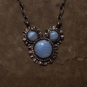 May include: A necklace featuring a large central round blue lace agate cabochon flanked by two smaller matching cabochons. The stones are set in copper with twisted wire and ruffled metal edges, suspended from a thick copper oval link chain.