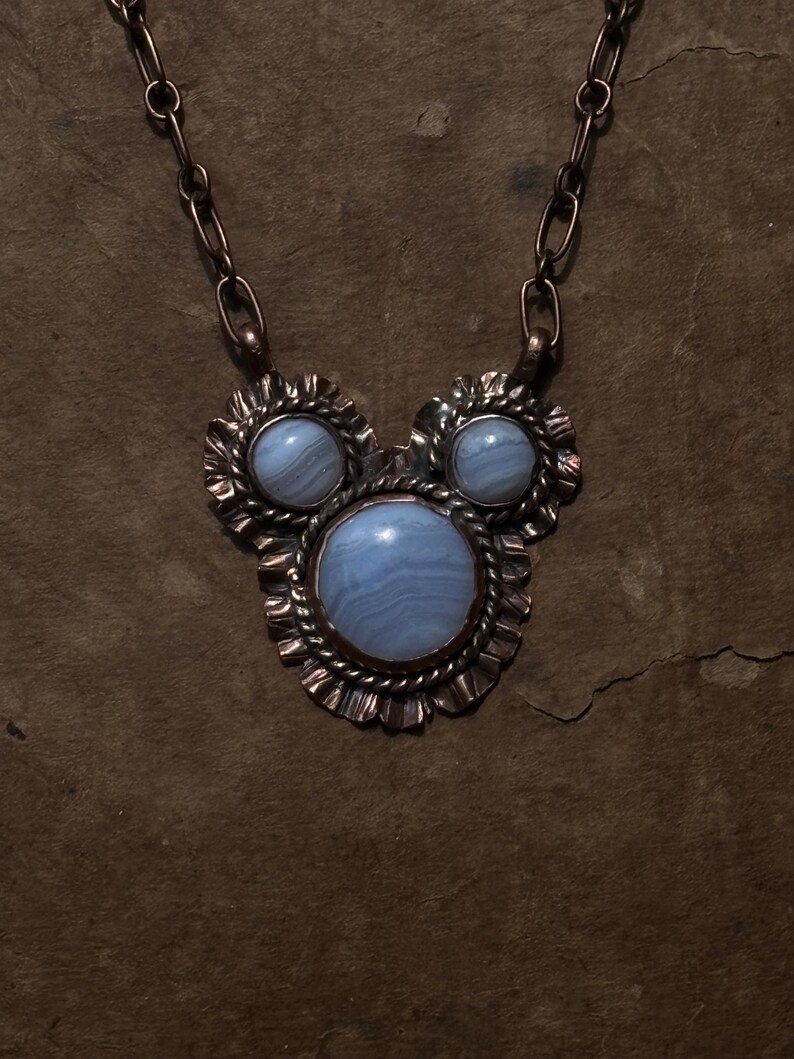 May include: A necklace featuring a large central round blue lace agate cabochon flanked by two smaller matching cabochons. The stones are set in copper with twisted wire and ruffled metal edges, suspended from a thick copper oval link chain.