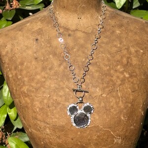 May include: A silver-toned necklace featuring a chain of interconnected circular links and a toggle clasp. The pendant consists of three dark, circular stones set in a silver frame, arranged in a shape resembling a stylized mouse head silhouette.