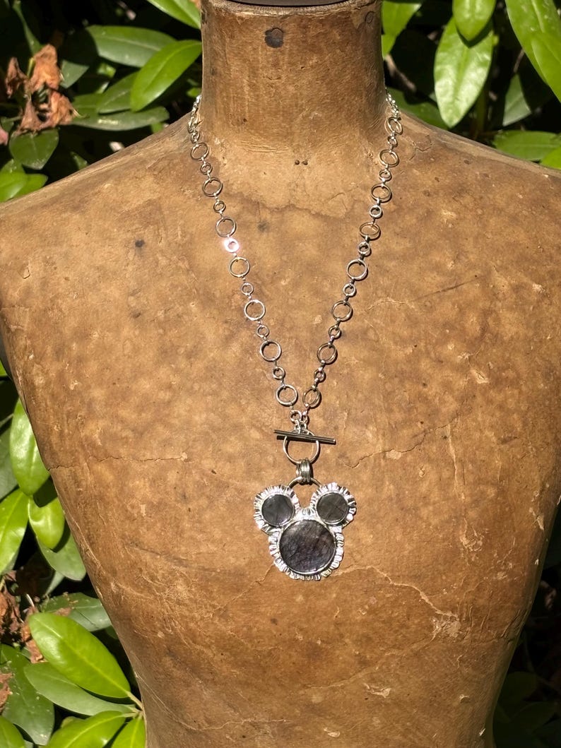 May include: A silver-toned necklace featuring a chain of interconnected circular links and a toggle clasp. The pendant consists of three dark, circular stones set in a silver frame, arranged in a shape resembling a stylized mouse head silhouette.