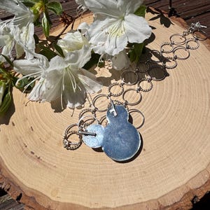 May include: A silver necklace with a blue pendant and two smaller silver circles. The necklace is displayed on a wooden surface with white flowers and green leaves. The pendant has a textured, light blue surface.