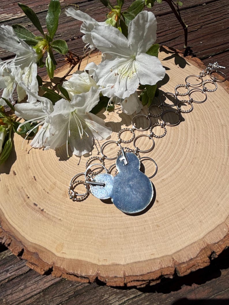 May include: A silver necklace with a blue pendant and two smaller silver circles. The necklace is displayed on a wooden surface with white flowers and green leaves. The pendant has a textured, light blue surface.