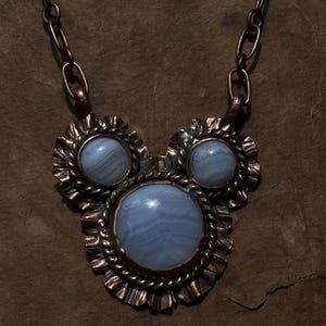 May include: A necklace featuring three round, light blue, banded agate cabochons set in dark, oxidized copper. The central stone is the largest, flanked by two smaller stones. Each stone is framed by a twisted wire border and a ruffled metal edge.