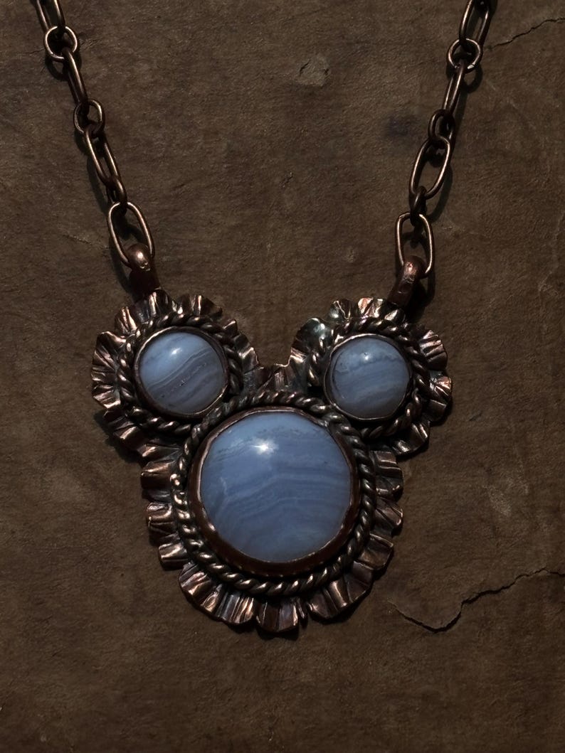 May include: A necklace featuring three round, light blue, banded agate cabochons set in dark, oxidized copper. The central stone is the largest, flanked by two smaller stones. Each stone is framed by a twisted wire border and a ruffled metal edge.