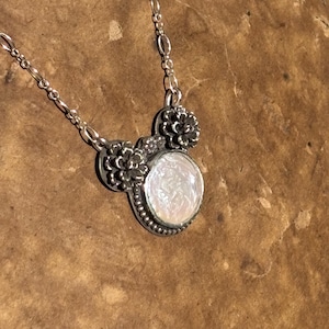 K&ouml;nnte beinhalten: Eine silberne Halskette mit einem runden Anh&auml;nger, der einen schimmernden wei&szlig;en Stein enth&auml;lt. Der Anh&auml;nger ist mit kleinen metallischen Blumenverzierungen und einem Perlenrand versehen und h&auml;ngt an einer feinen Silberkette mit ovalen Gliedern.