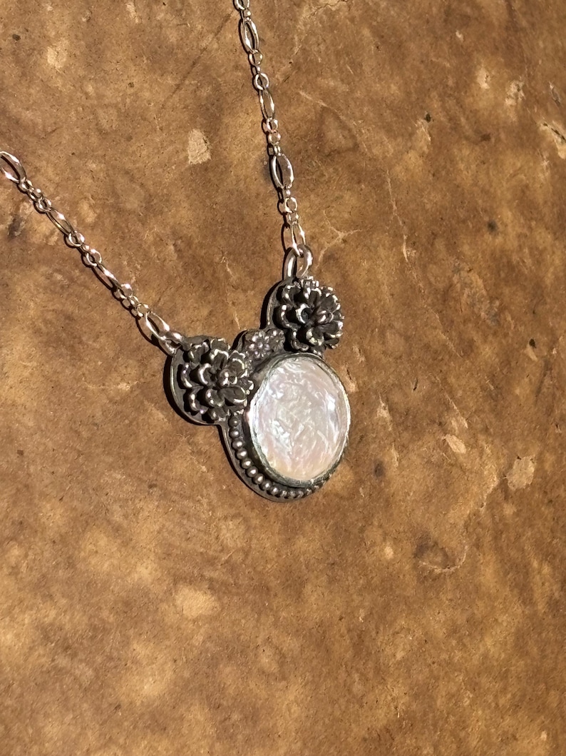 K&ouml;nnte beinhalten: Eine silberne Halskette mit einem runden Anh&auml;nger, der einen schimmernden wei&szlig;en Stein enth&auml;lt. Der Anh&auml;nger ist mit kleinen metallischen Blumenverzierungen und einem Perlenrand versehen und h&auml;ngt an einer feinen Silberkette mit ovalen Gliedern.