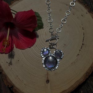 May include: A silver-toned necklace featuring a pendant with three circular iridescent stones set in a ruffled metal frame. The pendant is attached to a toggle clasp and a chain composed of interconnected silver rings of varying sizes.