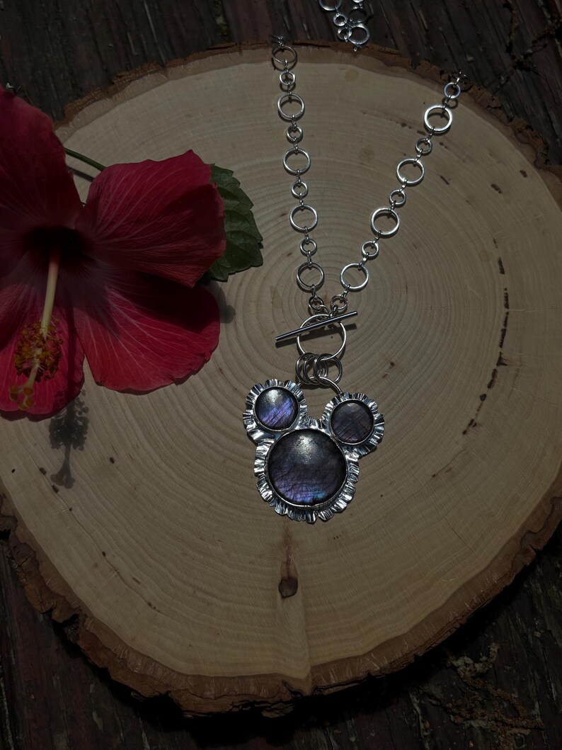 May include: A silver-toned necklace featuring a pendant with three circular iridescent stones set in a ruffled metal frame. The pendant is attached to a toggle clasp and a chain composed of interconnected silver rings of varying sizes.