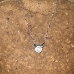 May include: A silver-toned necklace featuring a delicate chain and a circular pendant. The pendant has a white, iridescent center piece framed by a metallic border with small floral-like accents at the top where it connects to the chain.