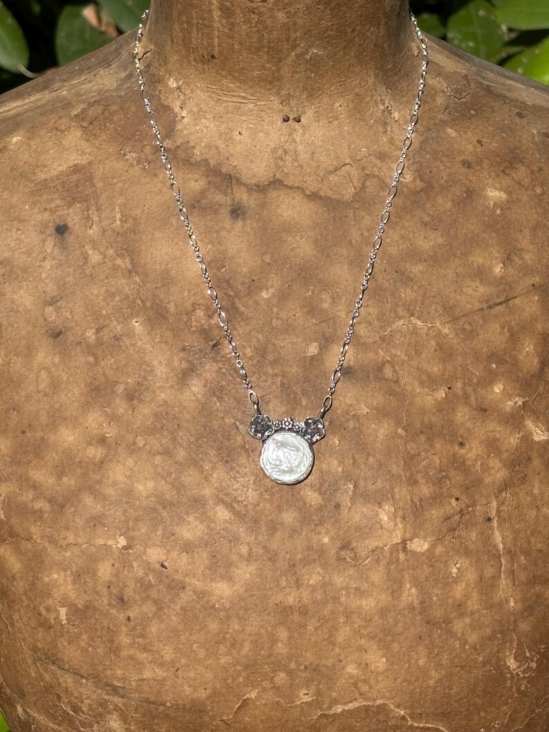 May include: A silver-toned necklace featuring a delicate chain and a circular pendant. The pendant has a white, iridescent center piece framed by a metallic border with small floral-like accents at the top where it connects to the chain.