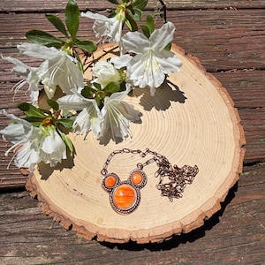 May include: A copper-toned necklace with an orange stone pendant, featuring a unique design. The necklace rests on a wooden slice, adorned with white flowers and green leaves. The necklace's chain is visible, adding to the overall aesthetic.