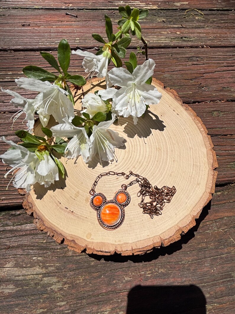 May include: A copper-toned necklace with an orange stone pendant, featuring a unique design. The necklace rests on a wooden slice, adorned with white flowers and green leaves. The necklace's chain is visible, adding to the overall aesthetic.