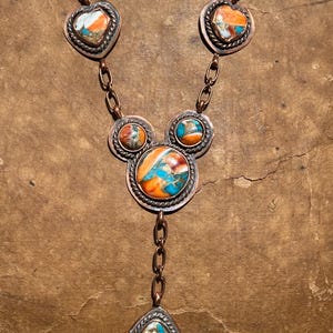 May include: A copper necklace featuring multiple stones with orange, white, and turquoise blue marbling. The design includes two heart-shaped settings at the top, a central cluster of three circular settings, and a single teardrop-shaped pendant at the base.