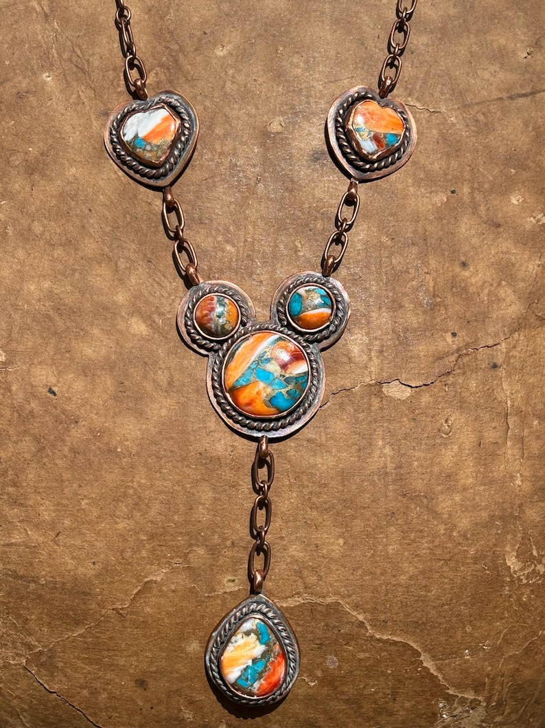 May include: A copper necklace featuring multiple stones with orange, white, and turquoise blue marbling. The design includes two heart-shaped settings at the top, a central cluster of three circular settings, and a single teardrop-shaped pendant at the base.