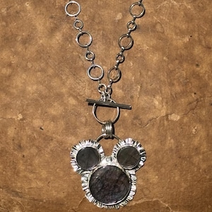 May include: A silver-toned necklace featuring a chain of interconnected circular links and a toggle clasp. The pendant consists of three dark, circular stones set in silver frames, arranged in a cluster resembling a three-circle silhouette.