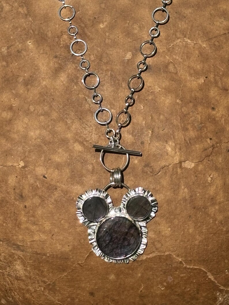 May include: A silver-toned necklace featuring a chain of interconnected circular links and a toggle clasp. The pendant consists of three dark, circular stones set in silver frames, arranged in a cluster resembling a three-circle silhouette.