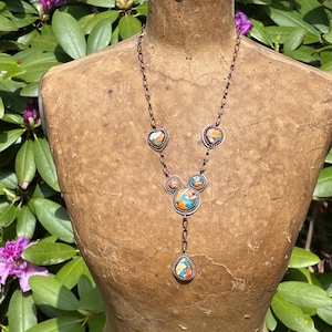 May include: A copper-toned necklace featuring a chain with multiple circular and heart-shaped settings. Each setting contains inlaid stone fragments in shades of orange, blue, and white. The design includes a central cluster and a teardrop pendant.