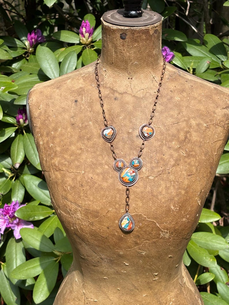 May include: A copper-toned necklace featuring a chain with multiple circular and heart-shaped settings. Each setting contains inlaid stone fragments in shades of orange, blue, and white. The design includes a central cluster and a teardrop pendant.