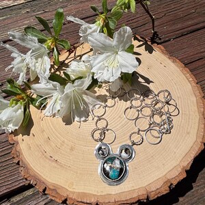 May include: A silver necklace with a Mickey Mouse-shaped pendant featuring a turquoise and white stone. The necklace is displayed on a wooden slice with white flowers and green leaves.