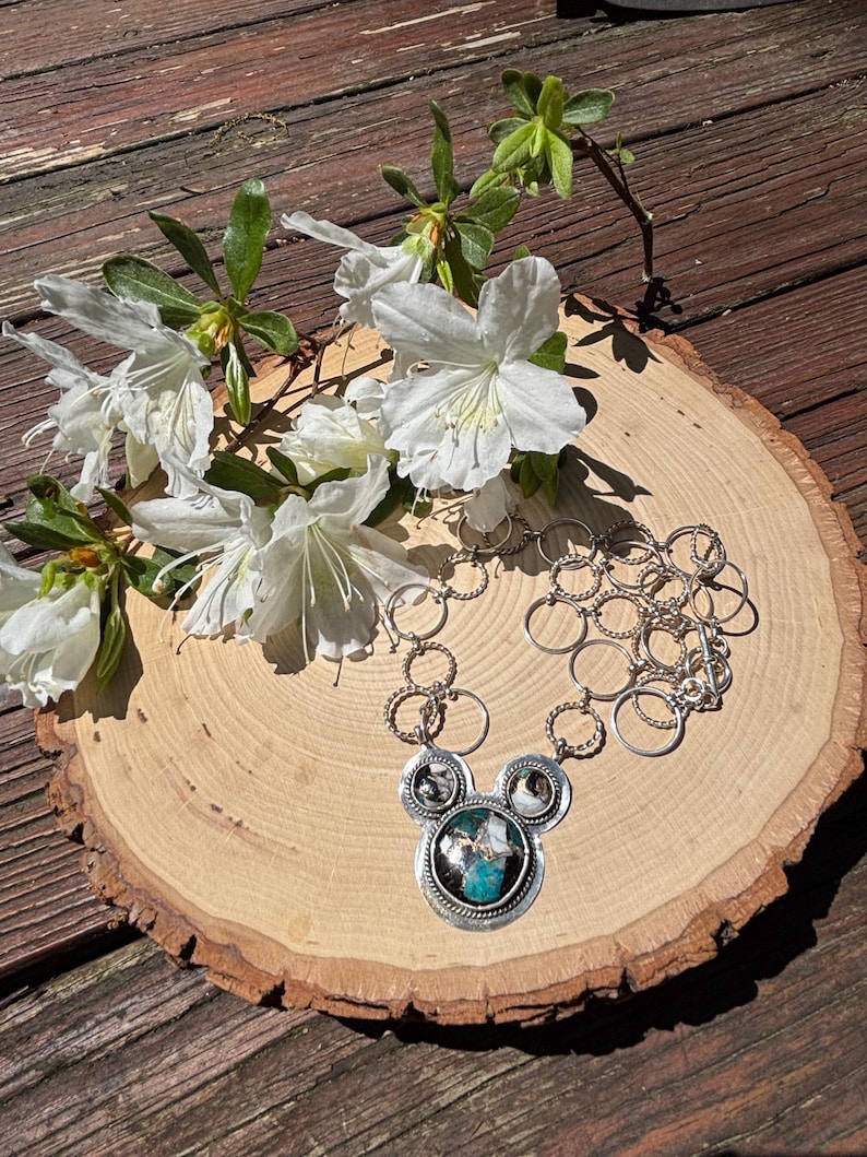 May include: A silver necklace with a Mickey Mouse-shaped pendant featuring a turquoise and white stone. The necklace is displayed on a wooden slice with white flowers and green leaves.