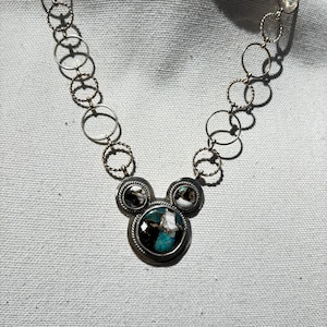 May include: Silver necklace with a pendant featuring three circular settings. The central setting has a turquoise stone with black and white details, and the side settings have black and white patterned stones. The chain is made of interconnected circles.