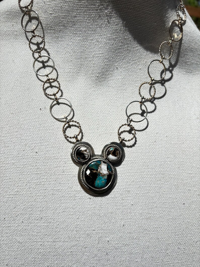 May include: Silver necklace with a pendant featuring three circular settings. The central setting has a turquoise stone with black and white details, and the side settings have black and white patterned stones. The chain is made of interconnected circles.