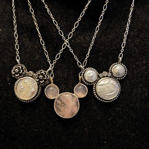 Puede incluir: Tres collares de plata con dise&ntilde;os de colgantes. Los colgantes presentan una piedra central grande, rodeada de piedras m&aacute;s peque&ntilde;as y detalles florales o circulares. Los collares cuelgan sobre un fondo oscuro.
