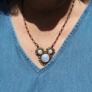 May include: A copper-toned metal necklace featuring a central pendant with three round, light blue, banded agate stones set in a decorative, textured frame. The pendant is attached to a matching copper-colored oval link chain.
