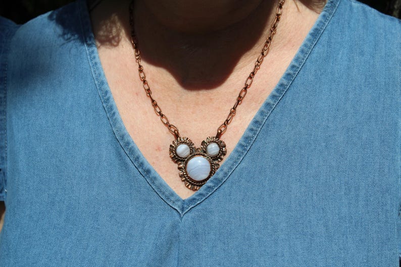 May include: A copper-toned metal necklace featuring a central pendant with three round, light blue, banded agate stones set in a decorative, textured frame. The pendant is attached to a matching copper-colored oval link chain.