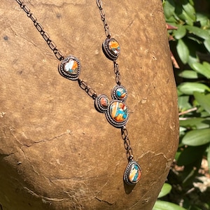 May include: A copper-toned metal necklace featuring a chain with multiple inlaid stone settings. The stones display a marbled pattern of orange, blue, and white. The central pendant consists of three circular settings arranged in a cluster.