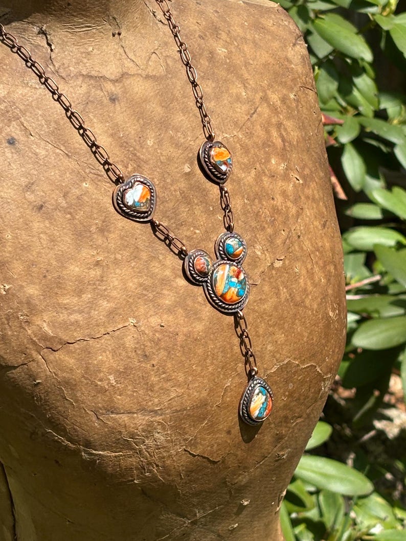 May include: A copper-toned metal necklace featuring a chain with multiple inlaid stone settings. The stones display a marbled pattern of orange, blue, and white. The central pendant consists of three circular settings arranged in a cluster.