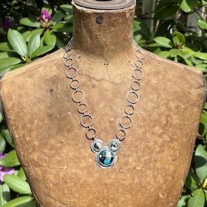 May include: Silver necklace with a Mickey Mouse-shaped pendant. The pendant has a turquoise and black stone centre, with two smaller stones either side. The necklace is displayed on a vintage mannequin.