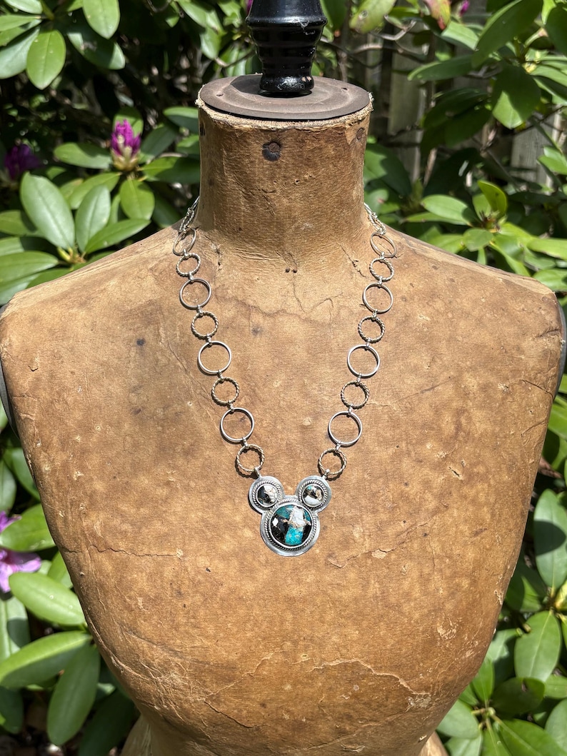 May include: Silver necklace with a Mickey Mouse-shaped pendant. The pendant has a turquoise and black stone centre, with two smaller stones either side. The necklace is displayed on a vintage mannequin.
