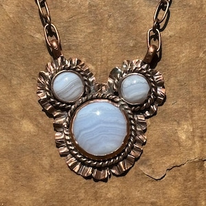 May include: A necklace featuring three round, light blue banded agate stones set in copper. The central stone is the largest, flanked by two smaller stones. Each stone is surrounded by a twisted copper wire border and a ruffled copper edge.