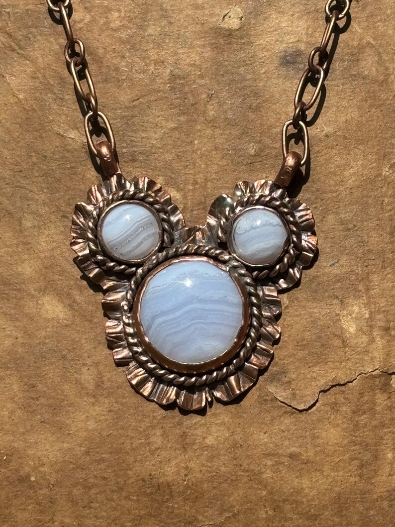 May include: A necklace featuring three round, light blue banded agate stones set in copper. The central stone is the largest, flanked by two smaller stones. Each stone is surrounded by a twisted copper wire border and a ruffled copper edge.