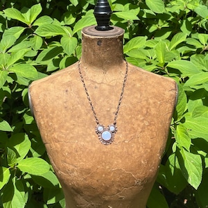 May include: A copper-toned necklace featuring a central pendant with three round, iridescent moonstone-like cabochons arranged in a cluster. The pendant is framed by a scalloped metal edge and hangs from a matching copper-toned oval link chain.