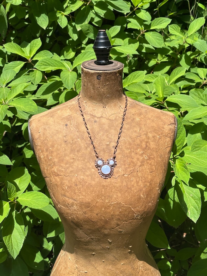 May include: A copper-toned necklace featuring a central pendant with three round, iridescent moonstone-like cabochons arranged in a cluster. The pendant is framed by a scalloped metal edge and hangs from a matching copper-toned oval link chain.
