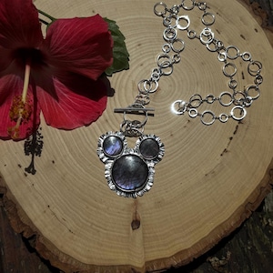 May include: A silver-toned necklace featuring a pendant with three circular, iridescent dark grey stones set in a scalloped metal frame. The pendant is attached to a chain composed of multiple interlocking silver-toned rings and a toggle clasp.