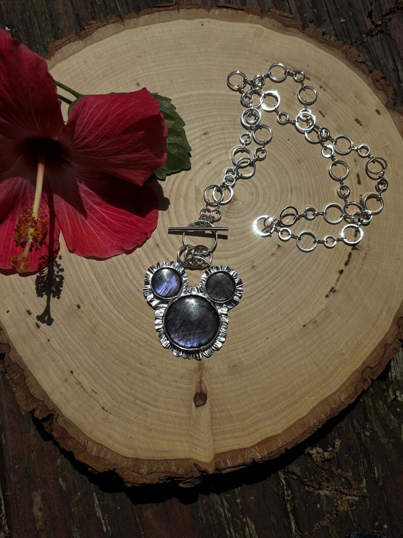 May include: A silver-toned necklace featuring a pendant with three circular, iridescent dark grey stones set in a scalloped metal frame. The pendant is attached to a chain composed of multiple interlocking silver-toned rings and a toggle clasp.