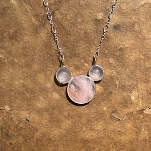 Puede incluir: Un collar de plata con tres piedras de cuarzo rosa. La piedra central, m&aacute;s grande, est&aacute; flanqueada por dos piedras m&aacute;s peque&ntilde;as. Las piedras tienen un suave tono rosado y est&aacute;n engastadas en biseles de plata. La cadena es una delicada cadena de plata.