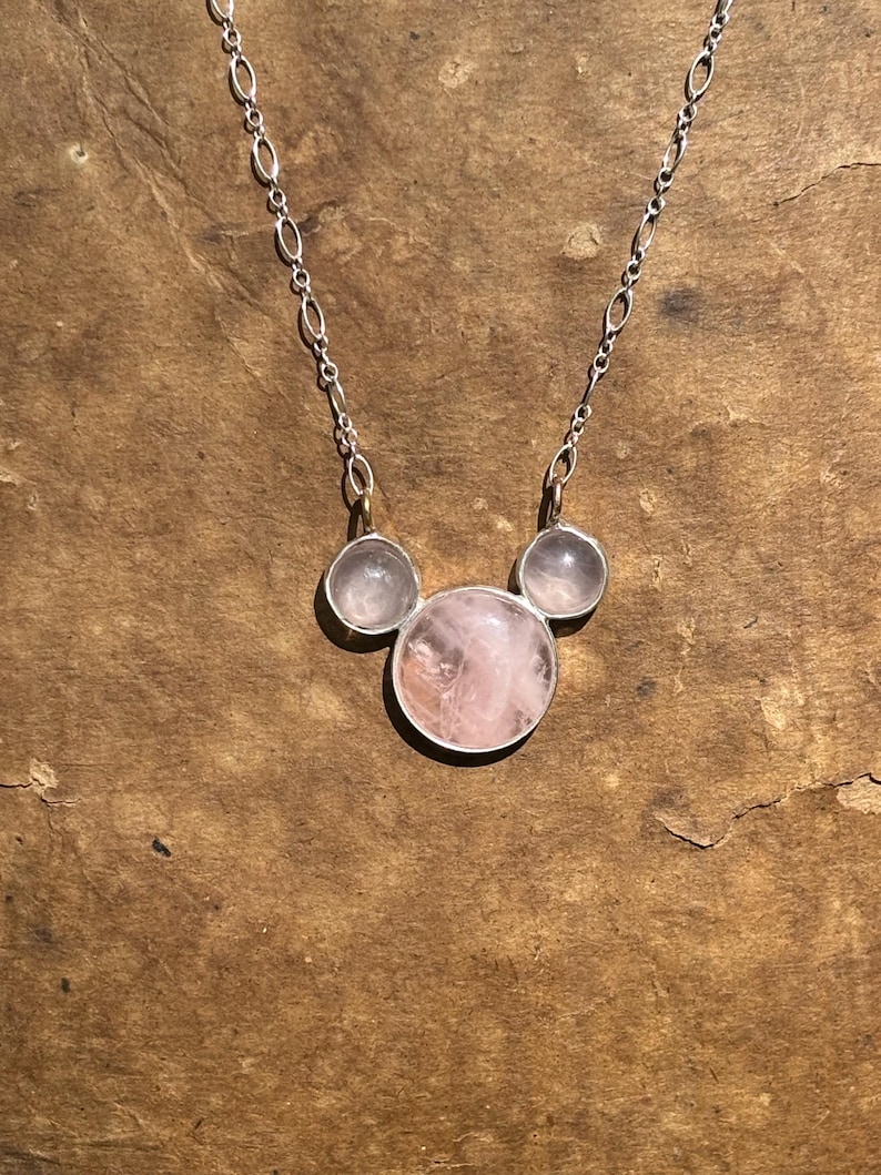 Puede incluir: Un collar de plata con tres piedras de cuarzo rosa. La piedra central, m&aacute;s grande, est&aacute; flanqueada por dos piedras m&aacute;s peque&ntilde;as. Las piedras tienen un suave tono rosado y est&aacute;n engastadas en biseles de plata. La cadena es una delicada cadena de plata.