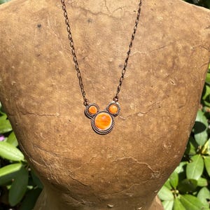 May include: A copper-toned necklace with three orange circular pendants. The central pendant is larger, with a textured border. The chain is a delicate link design. The necklace is displayed on a vintage mannequin.