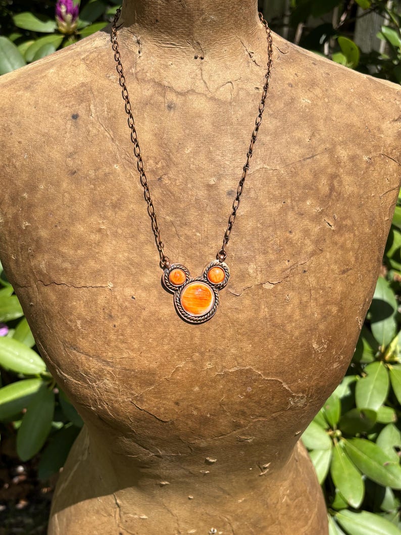 May include: A copper-toned necklace with three orange circular pendants. The central pendant is larger, with a textured border. The chain is a delicate link design. The necklace is displayed on a vintage mannequin.