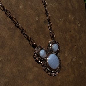 May include: A necklace featuring a copper chain and a pendant with three oval blue lace agate stones set in textured copper. The central stone is the largest, flanked by two smaller stones, all surrounded by a scalloped and twisted metal border.