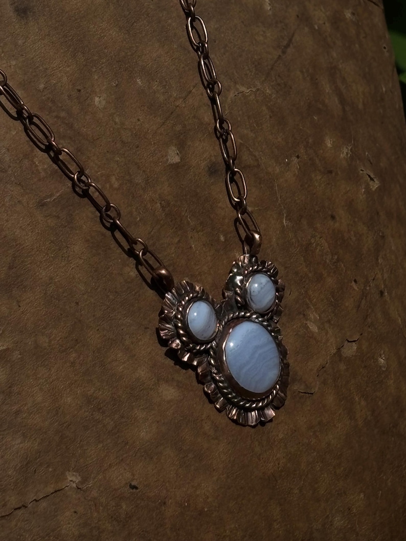 May include: A necklace featuring a copper chain and a pendant with three oval blue lace agate stones set in textured copper. The central stone is the largest, flanked by two smaller stones, all surrounded by a scalloped and twisted metal border.