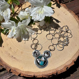 May include: A silver necklace with a pendant featuring a turquoise and black stone, with smaller stones. The necklace is displayed on a wooden slice with white flowers.