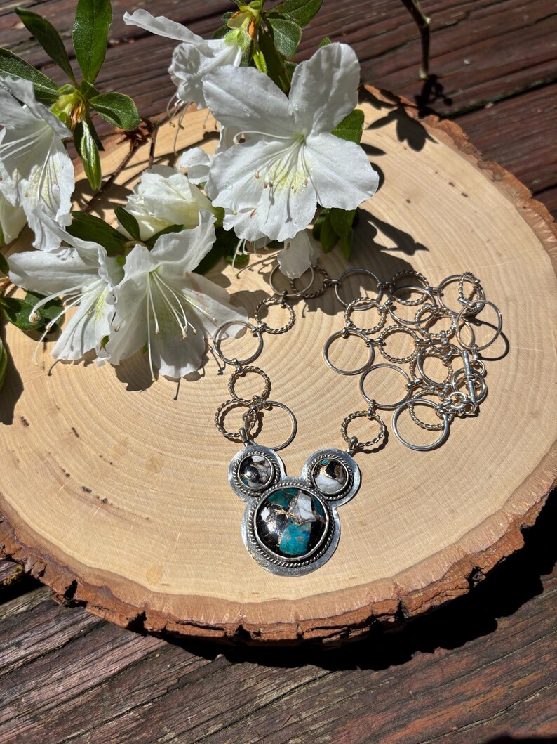 May include: A silver necklace with a pendant featuring a turquoise and black stone, with smaller stones. The necklace is displayed on a wooden slice with white flowers.