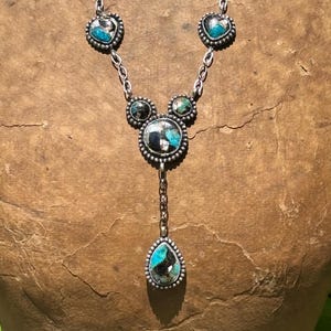 May include: A silver necklace featuring five bezel-set stones with turquoise and black matrix patterns. The design includes two heart-shaped stones, two small circular stones, one large central circular stone, and a dangling teardrop-shaped stone.
